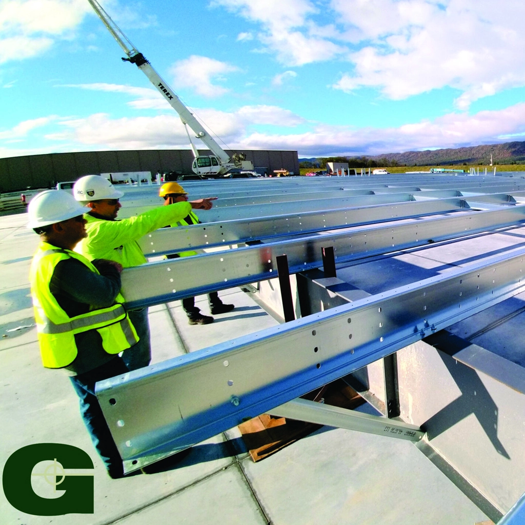 Construction workers in hard hats and safety vests assemble metal beams on a building roof under a clear blue sky