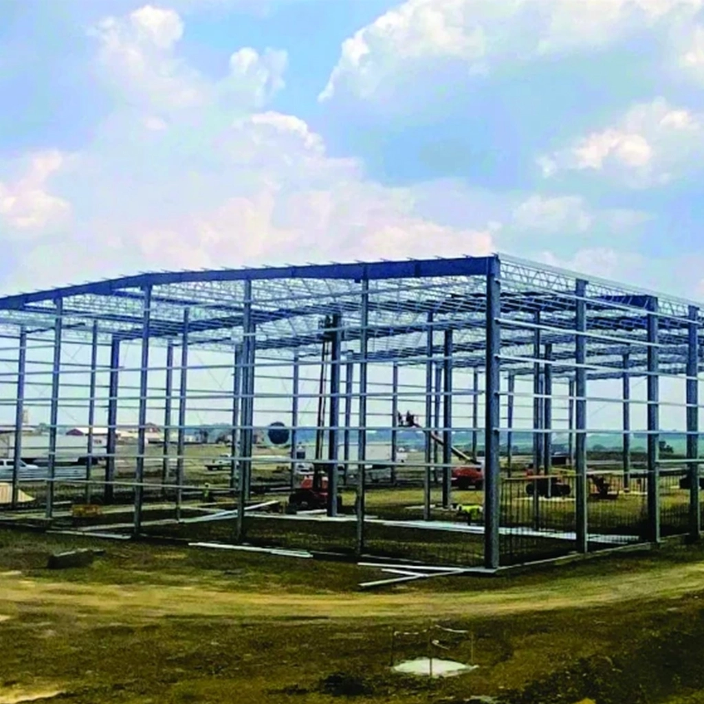 A partially constructed metal building on a construction site, with machinery and a crane under a cloudy sky. 