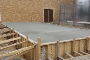 Freshly poured concrete slab surrounded by wooden formwork, with a brick wall and construction fencing in the background