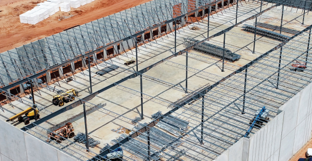 Construction workers in hard hats and safety vests assemble metal beams on a building roof under a clear blue sky